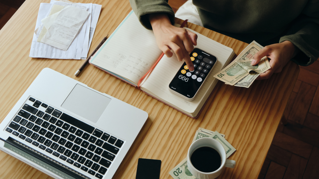 Business owner calculating finances with smartphone calculator, notebook, and U.S. currency on a desk.