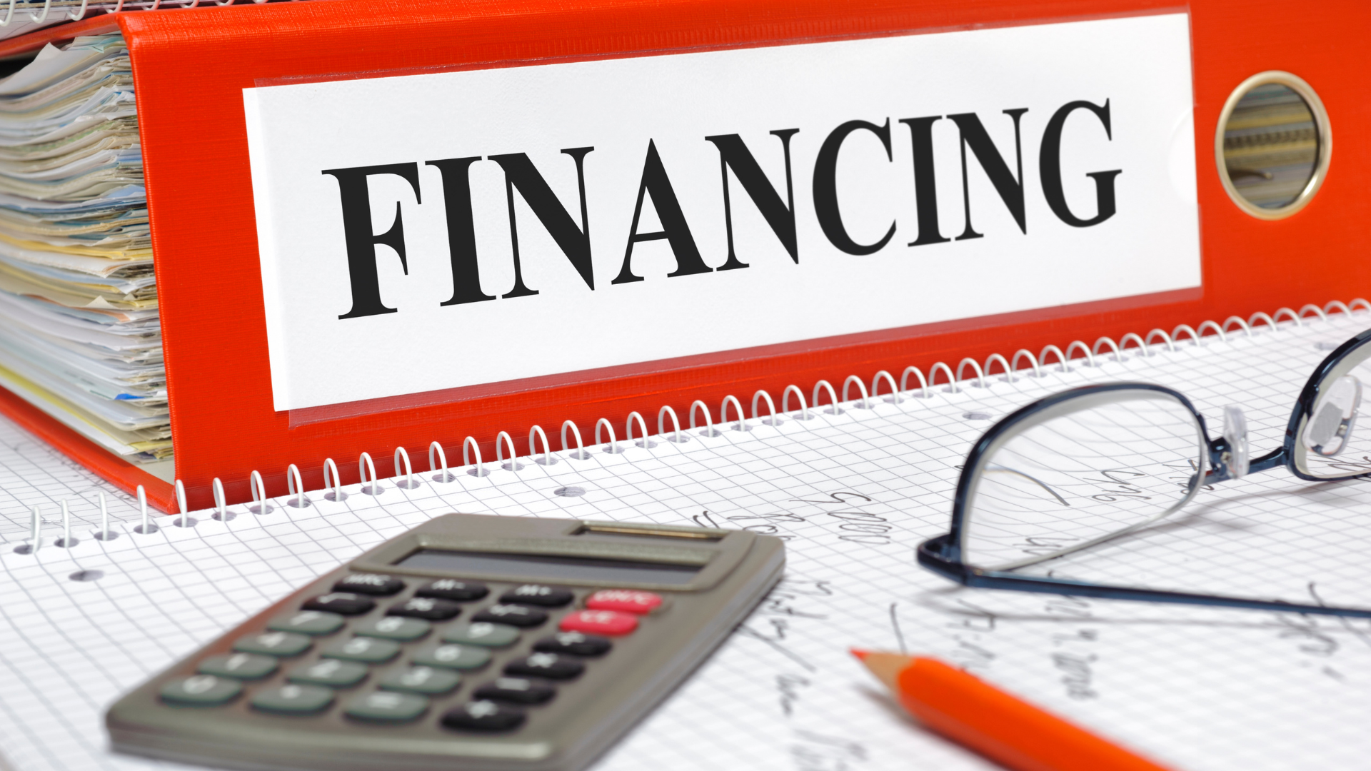 Close-up of a red binder labeled "FINANCING" with a calculator and eyeglasses on financial paperwork.
