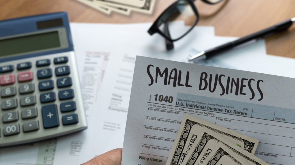 Small Business Loans header on a U.S. Income Tax form (1040) is visible, along with a calculator, a pen, and a stack of U.S. dollar bills. The image symbolizes the financial and tax-related aspects of managing a small business and applying for loans